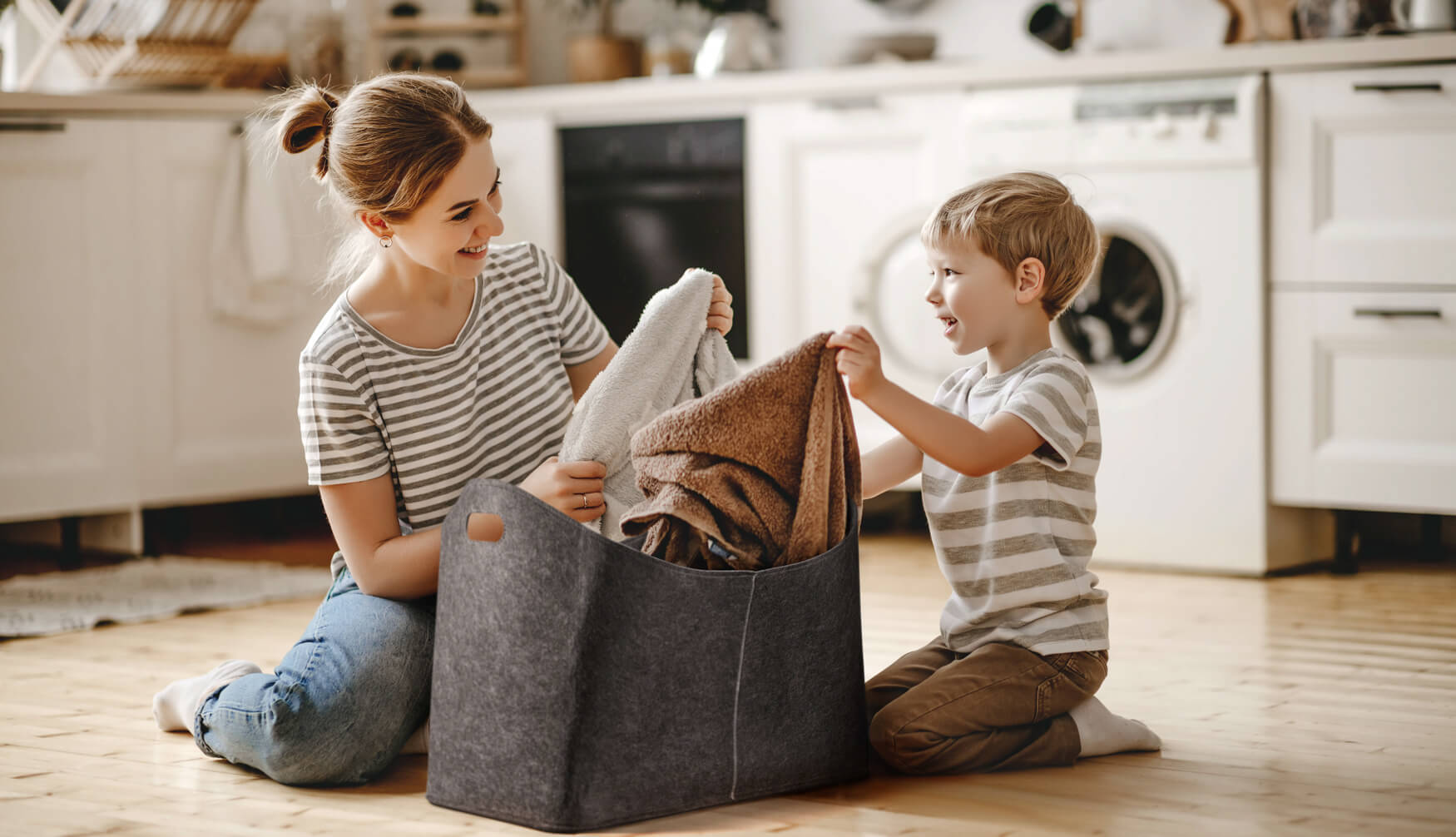 Ein herzerwärmendes Foto zeigt eine Mutter und ihr Kind neben einem Filzkorb mit Schmutzwäsche. Die Szene strahlt Geborgenheit und Alltagsleben aus, perfekt für Lifestyle-Fotografie und die Darstellung von Familienleben.