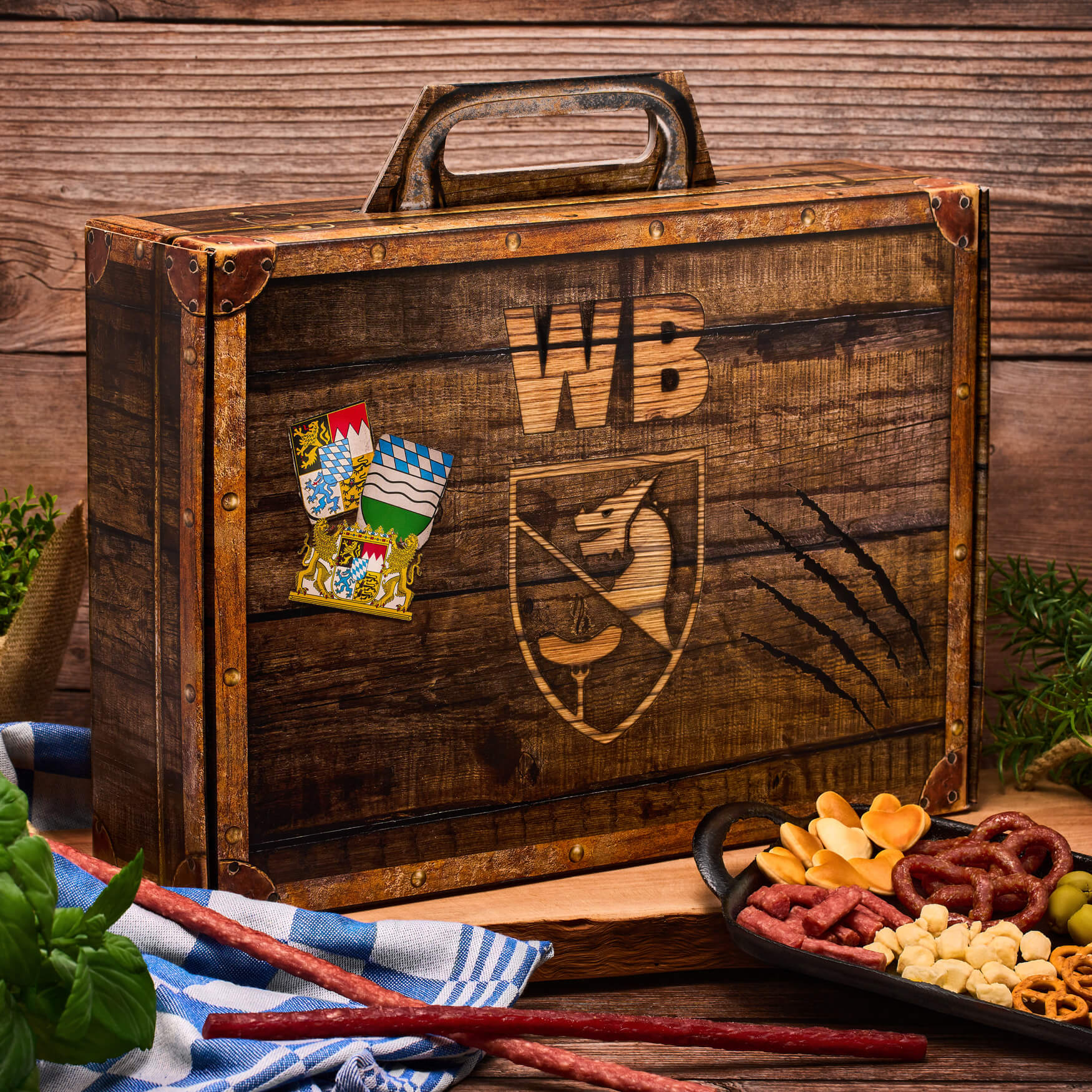 Geschenkverpackung: Koffer gefüllt mit Salami und Snacks auf rustikalem Holzhintergrund.