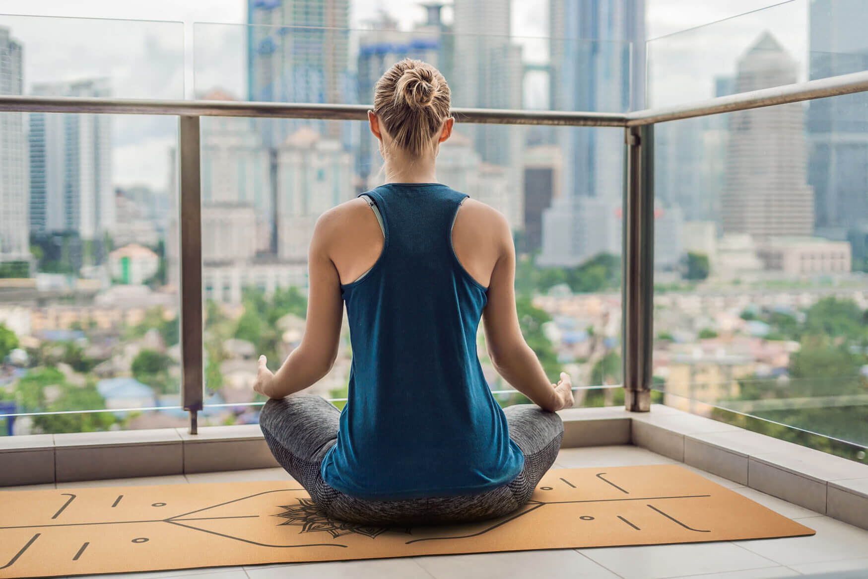 Ein beeindruckendes Lifestyle-Werbefoto einer Frau, die auf einer Yogamatte sitzt und die Skyline von einem Balkon eines Hochhauses aus betrachtet. Unsere Produktfotografie fängt die Ruhe und Gelassenheit dieser Szene ein, während die Frau inmitten der Großstadt ihre innere Balance findet und sich auf das Wohlbefinden konzentriert.