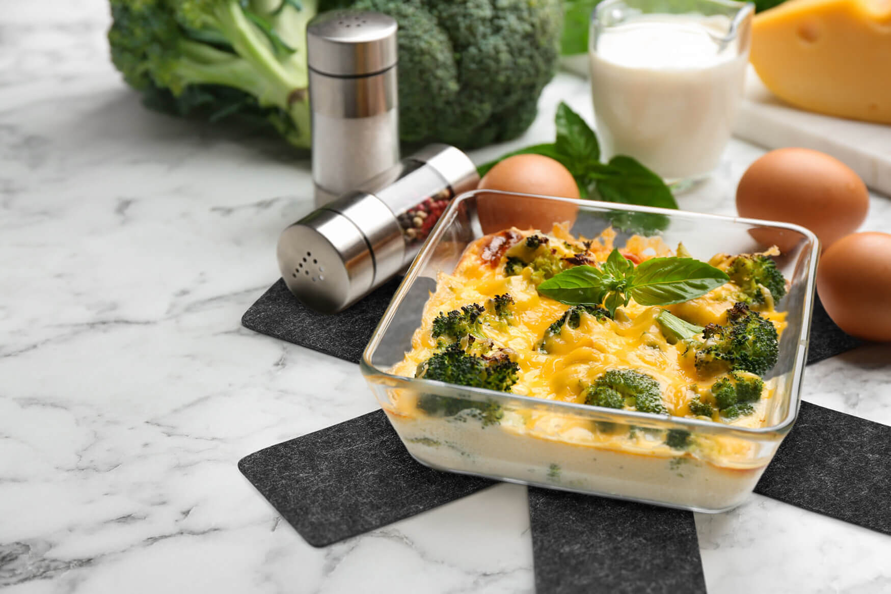 Eine ansprechende Lifestyle-Fotografie eines Brokkoliauflaufs auf einer Marmorarbeitsplatte, platziert auf einem Filzuntersetzer. Im Hintergrund sind dekorative Salz- und Pfefferstreuer, Eier, Milch, Käse sowie Brokkoli und Basilikum zu sehen. Unsere Produktfotografie fängt die Kulinarik und Eleganz dieses Gerichts ein und lädt den Betrachter ein, den Genuss der Zubereitung zu erleben.