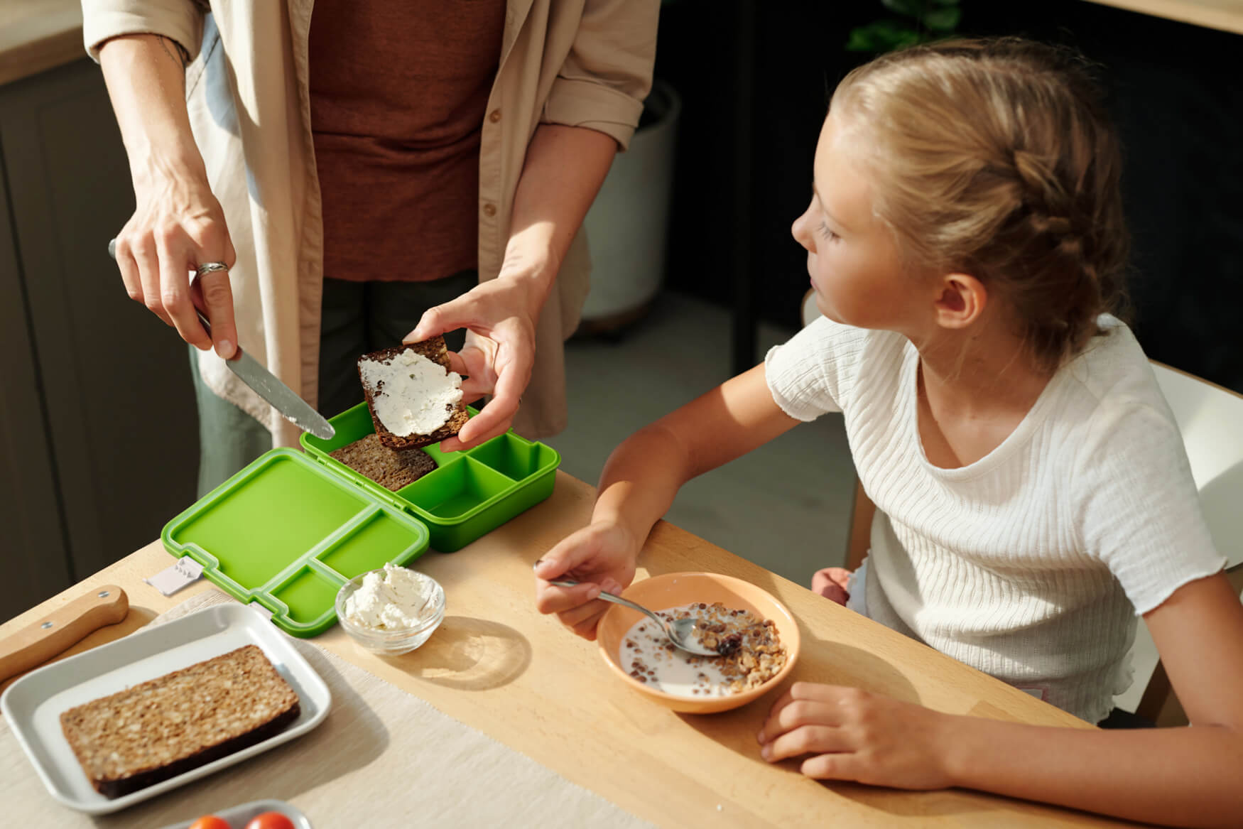 Eine Mutter bereitet liebevoll das Pausenbrot für ihre Tochter vor und packt es in die beworbene Lunchbox, während die Tochter frühstückt. Unser Foto vermittelt Geborgenheit und Fürsorge, während es gleichzeitig die Funktionalität und Qualität der Lunchbox hervorhebt. Durch solche emotionalen und ansprechenden Bilder helfen wir unseren Kunden, ihre Produkte erfolgreich zu präsentieren und Kunden zu überzeugen