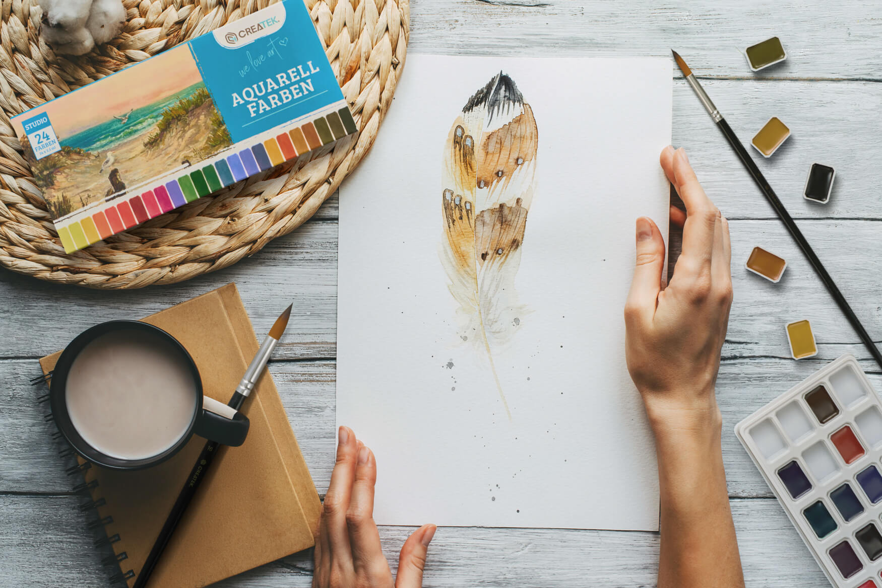 Ein Aquarellmalkasten, auf einem charmanten Landhaustisch drapiert, bildet die Kulisse für eine inspirierte Künstlerin, die mit zarter Hand eine Feder malt. Diese Szene vermittelt Kreativität, Leidenschaft und Kunstfertigkeit und weckt damit Emotionen, die potenzielle Kunden ansprechen und zu einem höheren Absatz führen können